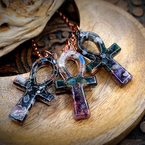 May include: Three ankh pendants with copper chains rest on a wooden plate. The ankhs are made of resin with embedded stones in shades of black, green, purple, and white. The pendants are a symbol of life.