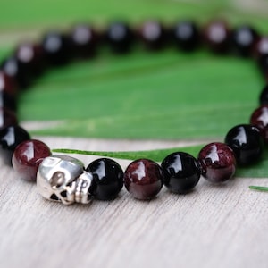 May include: A black and red beaded bracelet with a silver skull charm. The bracelet is made of round beads and is on a green leafy background.