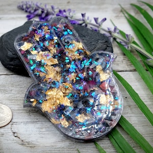 May include: A clear resin Hamsa hand charm with gold and iridescent glitter embedded inside. The Hamsa hand is a symbol of protection and good luck.