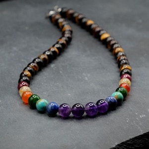 May include: A beaded necklace featuring a long strand of brown and gold tiger eye beads that transition into a vibrant sequence of multi-colored gemstone beads, including purple amethyst, blue lapis, turquoise, green jade, orange carnelian, and yellow citrine.
