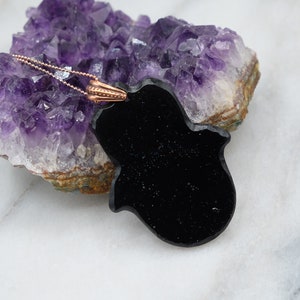 May include: Black onyx hamsa hand pendant necklace with a rose gold chain. The pendant is resting on a cluster of amethyst crystals.