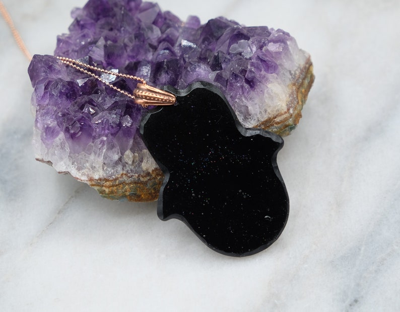 May include: Black onyx hamsa hand pendant necklace with a rose gold chain. The pendant is resting on a cluster of amethyst crystals.