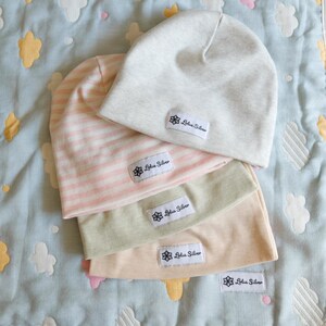 May include: Four soft beanies in light colors are stacked on a patterned blanket. The beanies are in shades of light gray, pink, green, and peach. Each beanie has a white label with the text "Lotus Silver".