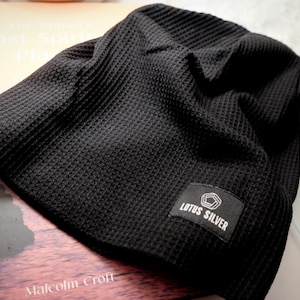May include: A black beanie with a textured waffle-knit pattern. The hat features a rectangular black fabric patch sewn onto the lower edge, displaying a white geometric pentagon logo above the white text that reads LOTUS SILVER.
