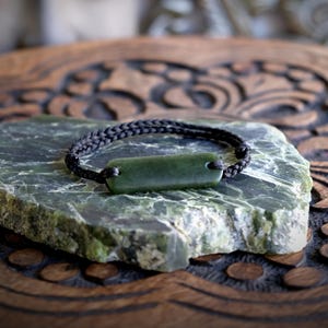 May include: A dark green jade bracelet with a rectangular jade stone pendant. The bracelet features a woven black cord. The jade stone is set on a rough, green stone surface, with a carved wooden background.