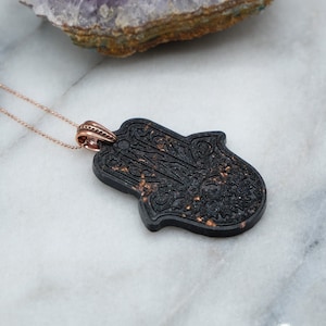 May include: A black hamsa hand pendant necklace with a rose gold chain. The hamsa hand is detailed with intricate patterns and has a speckled texture.