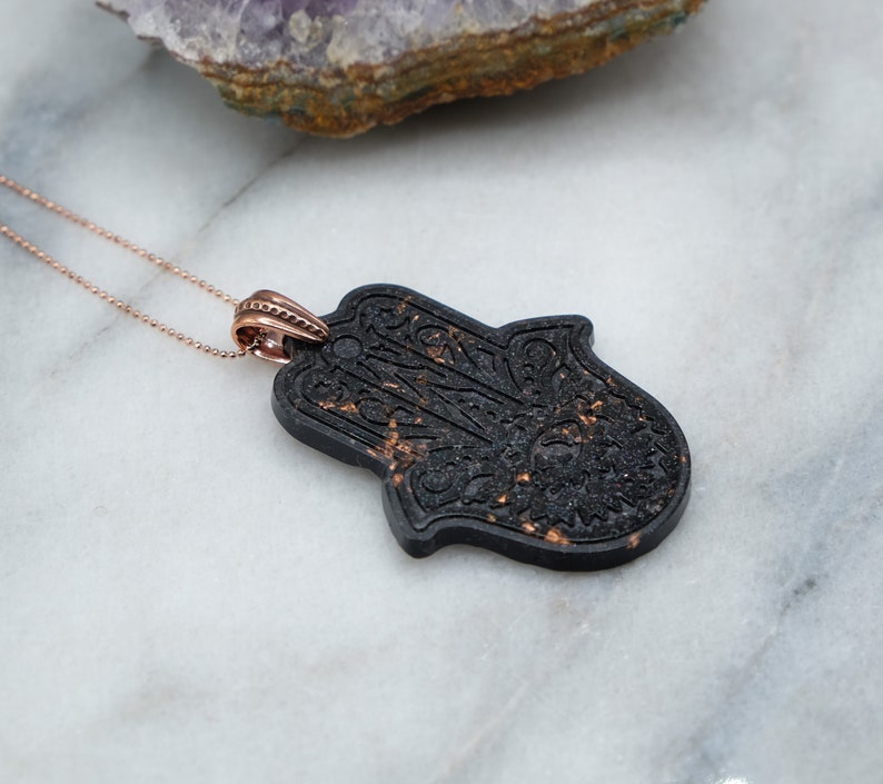 May include: A black hamsa hand pendant necklace with a rose gold chain. The hamsa hand is detailed with intricate patterns and has a speckled texture.