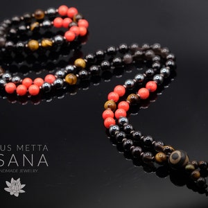 May include: A multi-strand mala necklace with black, brown, and orange beads. The beads are interspersed with coral-colored beads. The necklace has a tassel with a large, brown bead at the end. The necklace is called "Lotus Metta Asana Handmade Jewelry".