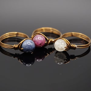 May include: Three gold wire wrapped rings with different colored stones. The first ring has a blue stone, the second ring has a pink stone, and the third ring has a white stone.