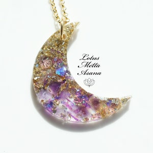 May include: A crescent moon pendant necklace with a gold chain. The moon pendant is filled with purple, gold, and iridescent glitter and crystals. The text "Lotus Metta Asana" is printed on the right side of the image.