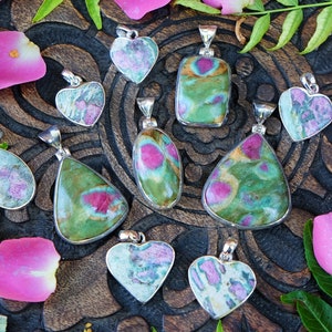 May include: A collection of 10 green and pink gemstone pendants with silver settings. The pendants are in various shapes, including hearts, teardrops, and ovals. The gemstones are a mix of green and pink, with some having a more pronounced pink hue.