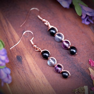 May include: A pair of dangle earrings with black, clear, and purple beads. The earrings are made with rose gold wire and have a simple hook closure.