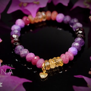 Peut inclure: Un bracelet en pierres précieuses colorées avec un dégradé de perles violettes, roses et orange. Le bracelet a un charme en forme de cœur en or et le texte "LOTUS METTA ASANA HANDMADE JEWELRY" est visible dans le coin inférieur gauche de l'image.