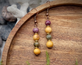 Peut inclure: Une paire de boucles d'oreilles pendantes avec trois perles chacune. La perle du haut est d'un violet foncé, la perle du milieu est d'un vert et d'un brun marbrés, et la perle du bas est d'un jaune clair. Les boucles d'oreilles sont suspendues à des crochets en cuivre.