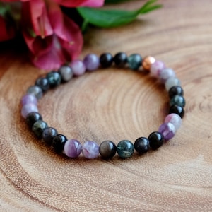May include: A beaded bracelet featuring a mix of black, purple, and green gemstones. The bracelet has a copper-colored accent bead. The beads are strung together to form a circular shape. The bracelet is set against a wooden surface.