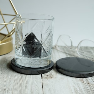 May include: A clear glass with a patterned design sits on a black coaster. The glass contains a black ice cube. A second black coaster is visible in the background.