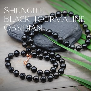 May include: A black beaded necklace made of shungite, black tourmaline, and obsidian stones. The necklace is lying on a wooden surface with green leaves and a black stone.
