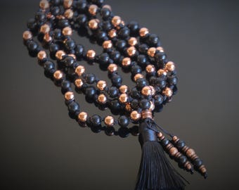 May include: A black and copper beaded mala necklace with a black tassel. The necklace is made of round beads and is strung on a black cord.