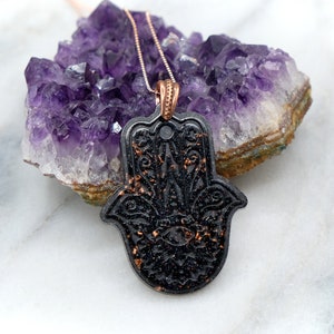 May include: A black hamsa hand pendant with gold glitter accents hangs from a rose gold chain. The pendant is resting on a cluster of amethyst crystals.