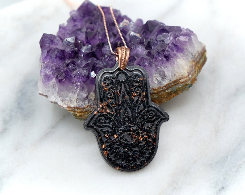May include: A black hamsa hand pendant with gold glitter accents hangs from a rose gold chain. The pendant is resting on a cluster of amethyst crystals.