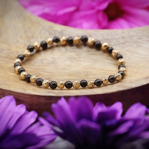May include: A black beaded bracelet with gold accents. The bracelet is made of small, round beads and is lying on a wooden surface.