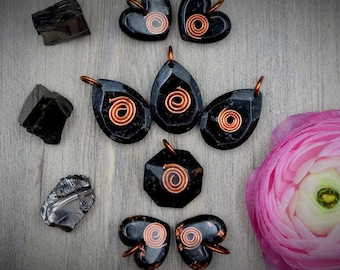 May include: A collection of black obsidian pendants with copper wire spirals. The pendants are in the shapes of hearts and teardrops. The pendants are arranged on a wooden surface with black obsidian stones and a pink flower.