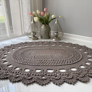 May include: A handcrafted, oval-shaped, crochet rug in a muted taupe color. The rug features an intricate openwork design around the edges, adding a delicate touch. The rug is placed on a white tiled floor, with a vase of flowers and decorative lanterns in the background.