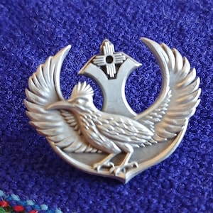 May include: A silver-colored metal pin featuring a roadrunner bird with outstretched wings, a stylized sun symbol, and a crescent-shaped design. The pin is set against a blue background.
