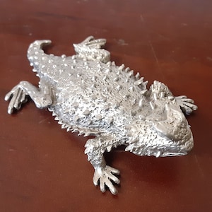 May include: A silver-colored metal sculpture of a horned lizard. The lizard is posed with its legs outstretched and its head tilted slightly to the side. The sculpture is detailed with textured scales and horns.