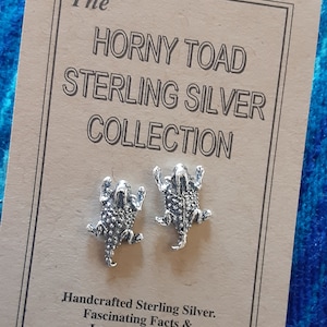 May include: A pair of sterling silver earrings featuring a horned toad design. The earrings are displayed on a card that reads "The Horny Toad Sterling Silver Collection." The card also states "Handcrafted Sterling Silver. Fascinating Facts & Legends on Back."