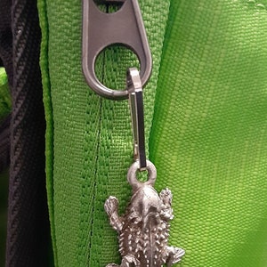 May include: A silver zipper charm shaped like a horned lizard. The charm is attached to a green zipper on a bag.