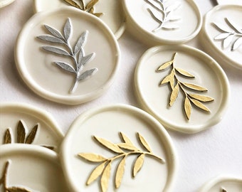 Puede incluir: Un conjunto de sellos de cera redondos con un diseño de hoja. Algunos sellos son blancos con diseños de hojas doradas, mientras que otros son beige con diseños de hojas plateadas.