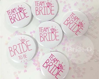 May include: Six white round badges with pink text and a pink diamond design. The badges read "Team Bride" and "Bride to Be".