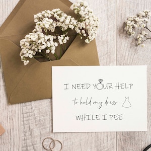 以下が含まれることがあります： 「I NEED YOUR HELP to hold my dress WHILE I PEE」というテキストが書かれた白いカードが表示されています。ダイヤモンドの指輪とドレスのグラフィックが含まれています。カードは、白い花が入った茶色の封筒の隣にあります。