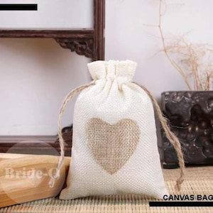 Puede incluir: Una bolsa de lona blanca con un diseño de corazón de arpillera marrón. La bolsa tiene un cierre de cordón y está hecha de tela natural.  CANVAS BAG está impreso en la esquina inferior derecha de la imagen.