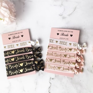 May include: Set of black, white and pink hair ties with gold glitter lettering. The hair ties say "Team Bride" and "Bride". The hair ties are packaged on a pink card with the text "Always look on the Bride side of life".