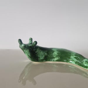 Pode incluir: Uma estatueta de lesma de cerâmica em tons de verde. A lesma tem um acabamento brilhante e está posicionada sobre uma superfície refletora. O corpo da estatueta é alongado, com um padrão texturizado, e tem dois pares de antenas.