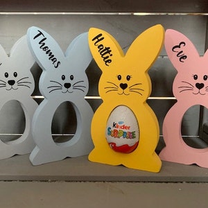 May include: Four wooden bunny-shaped Easter egg holders, painted in blue, yellow, pink, and light blue. Each bunny has a name written on it: Matthew, Thomas, Hattie, and Eve. One bunny has a Kinder Surprise egg inside.