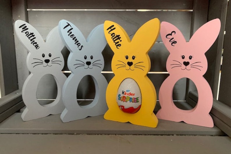 May include: Four wooden bunny-shaped Easter egg holders, painted in blue, yellow, pink, and light blue. Each bunny has a name written on it: Matthew, Thomas, Hattie, and Eve. One bunny has a Kinder Surprise egg inside.