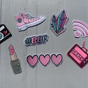 May include: A set of iron-on patches featuring a pink camera, a pink and white rainbow, a pink lipstick, a pink and white heart, a pink and white crystal, a pink and white shoe, a pink and white TV with the word "CRAZY" on it, and a pink and white patch with the text "#BBF".