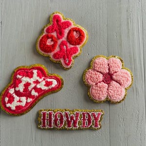 May include: Four embroidered patches in pink, red, and gold. Designs include an XOXO, a cowboy hat, a flower, and the word "HOWDY". The patches have a textured, raised appearance and are outlined in gold.
