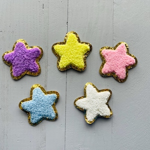 May include: Five fuzzy star-shaped embellishments with gold glitter trim. The stars are in shades of yellow, pink, purple, blue, and white.