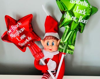 May include: A red and white Elf doll holding two star-shaped balloons. One balloon is red with the text "I'm Back Meghan Love Sparkle". The other is green with the text "Isabelle Jack We're Back". The elf is wearing a red and white outfit.