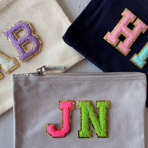 May include: Three zippered pouches with different coloured felt letters. The first pouch is beige with white and gold letters "AB". The second pouch is navy blue with pink and gold letters "HM". The third pouch is grey with pink and gold letters "JN".