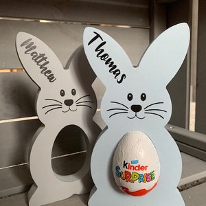 May include: Two wooden bunny-shaped Easter egg holders, one light blue and one light gray. The blue bunny has a Kinder Surprise egg in it. The bunnies have the names "Thomas" and "Matthew" written on them.