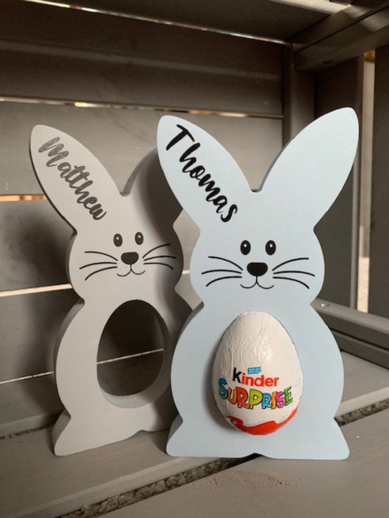 May include: Two wooden bunny-shaped Easter egg holders, one light blue and one light gray. The blue bunny has a Kinder Surprise egg in it. The bunnies have the names "Thomas" and "Matthew" written on them.