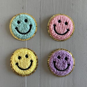 May include: Four embroidered smiley face patches with different coloured fuzzy backgrounds. The patches are light blue, pink, yellow, and purple.