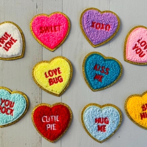 May include: A selection of heart-shaped patches in various colours, each with a gold glitter border. The patches display phrases like "True Love," "Sweet," "XOXO," "Love You," "Love Bug," "Kiss Me," "You Rock," "Cutie Pie," "Hug Me," and "Be Mine."