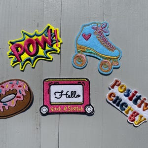 May include: Five embroidered iron-on patches. The patches are colourful and feature a donut, a roller skate, a drawing of an Etch A Sketch, a word bubble with the word "POW!" and a rainbow coloured word that says "Positive Energy".
