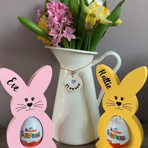 May include: Two wooden bunny shaped Easter egg holders, one pink and one yellow, with the names "Eve" and "Hattie" written on them. Each bunny is holding a Kinder Surprise chocolate egg. A white ceramic pitcher with a blue heart charm that says "Flowers" is in the background.